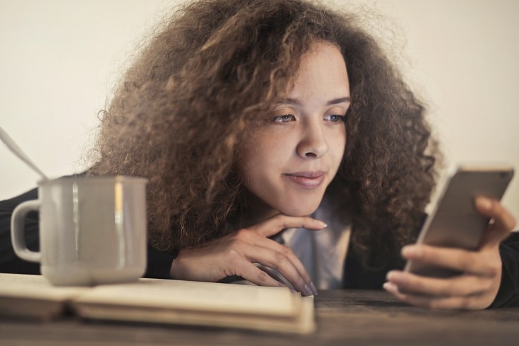 Junge Frau sitzt entspannt an einem Tisch und schaut zufrieden auf ein <Smartphone